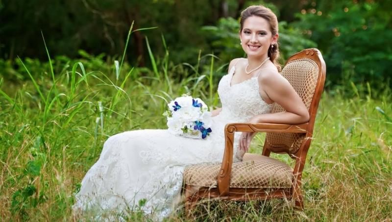 Bride in white dress sitting on wicker chair in a grassy field, holding a bouquet and smiling
