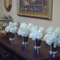 White flowers in silver vases arranged on a dark wooden dresser beneath framed wall art.