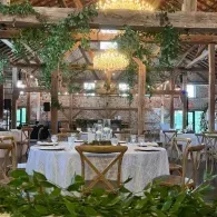 Rustic banquet hall with round white-clothed tables, wooden chairs, greenery, and hanging lights