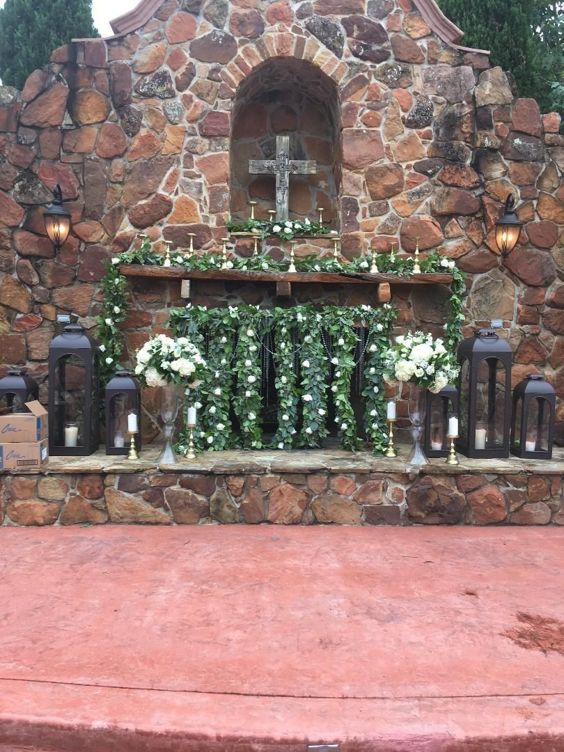 Stone grotto altar with white flowers and lanterns in front of a cross, set against a rock wall.