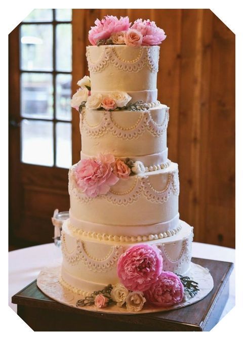 Four-tier white wedding cake with pink flowers and lace piping on a table