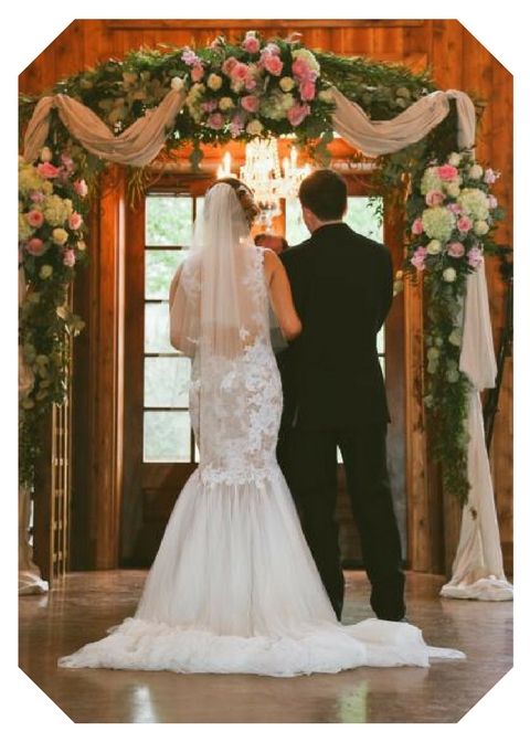 Bride and groom stand beneath a floral arch in a rustic, flower-filled wedding venue.