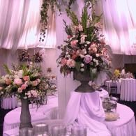 Floral centerpiece on a draped reception table under soft pink lighting, with matching bouquets nearby