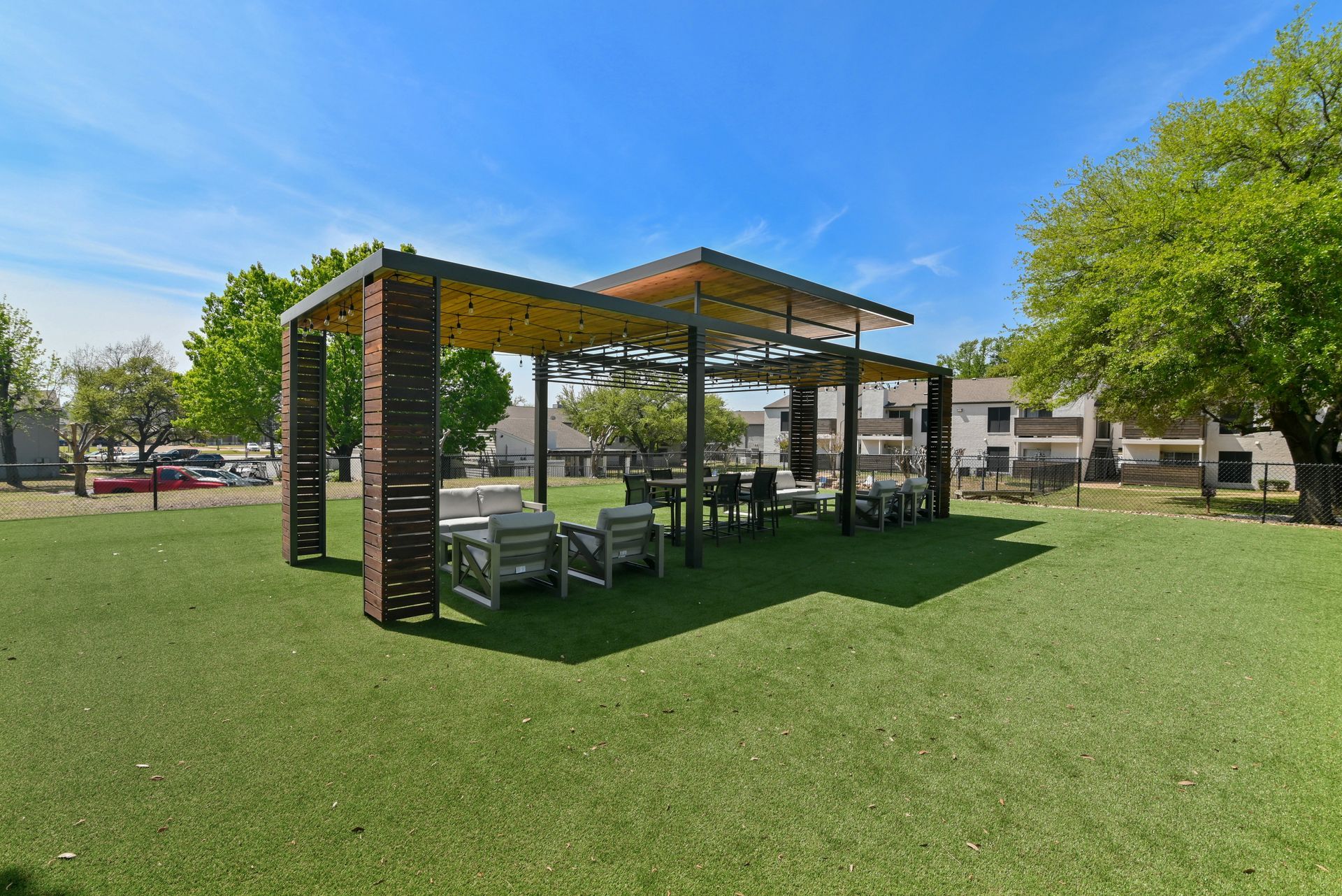 Outdoor seating area with pergola, tables, and chairs on green turf under a blue sky.