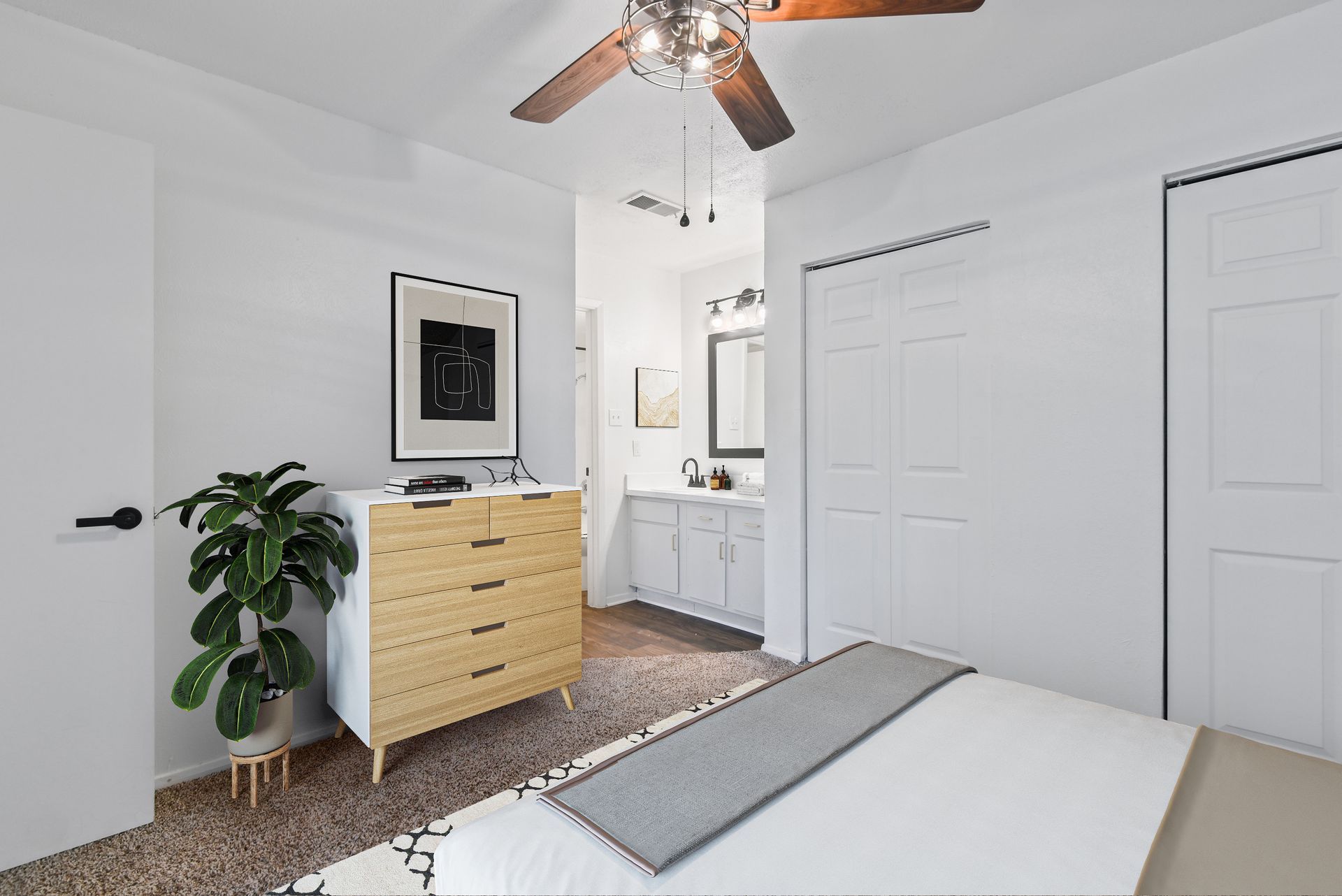 Bedroom with white walls, a dresser, a plant, a bed, and a bathroom visible in the background.