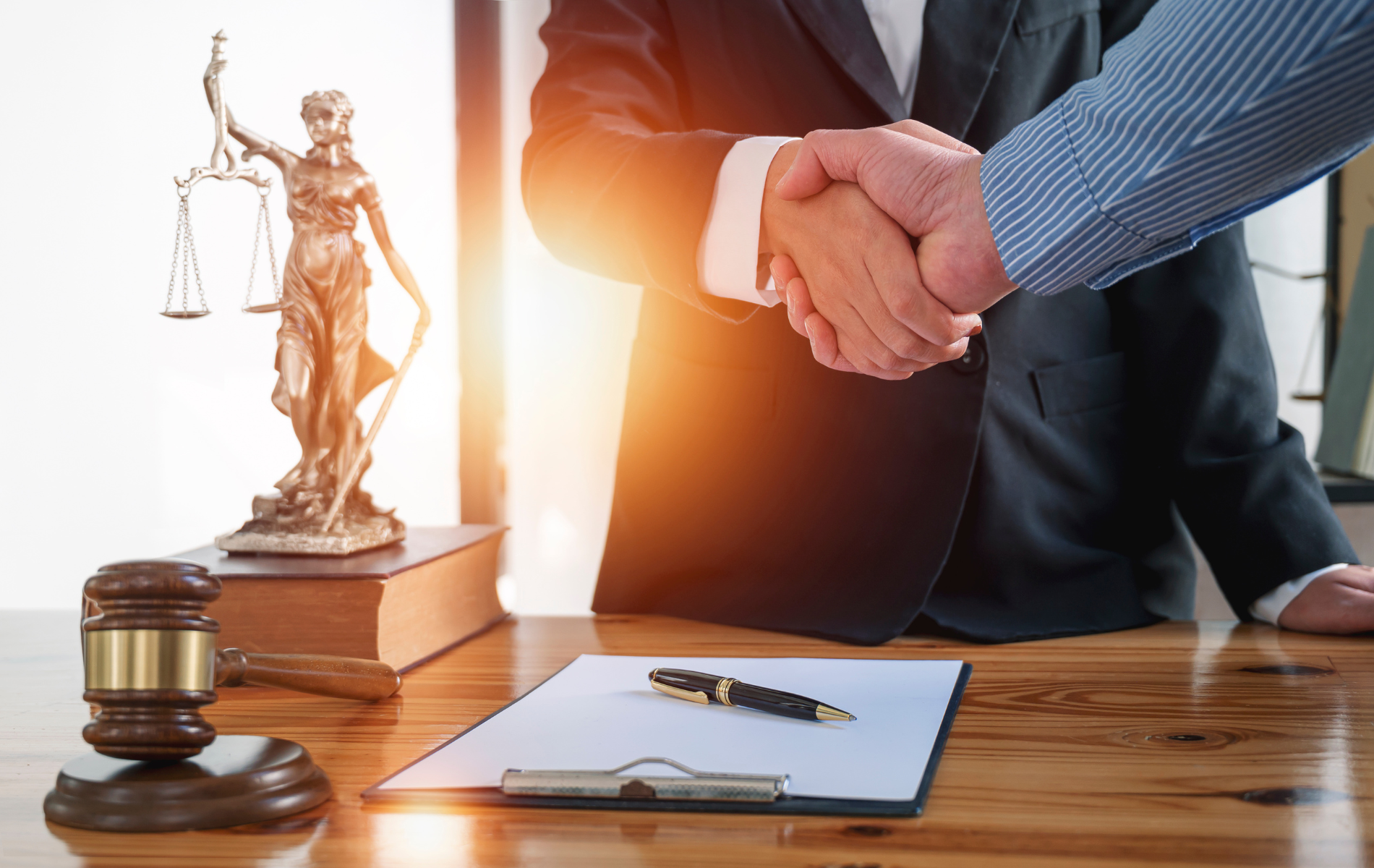 Handshake over lawyer’s desk with gavel, scales of justice, and signed document folder