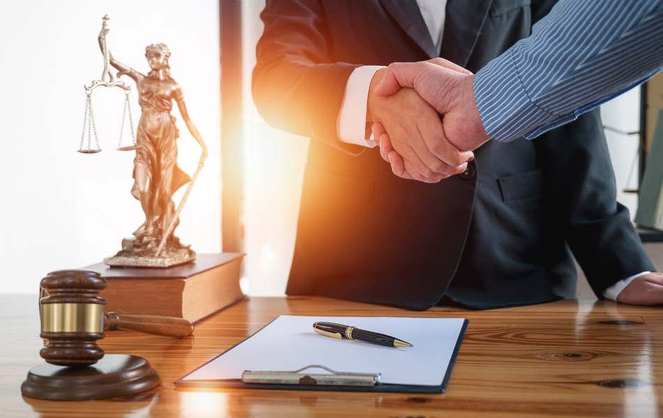 Handshake over lawyer’s desk with gavel, scales of justice, and signed document folder