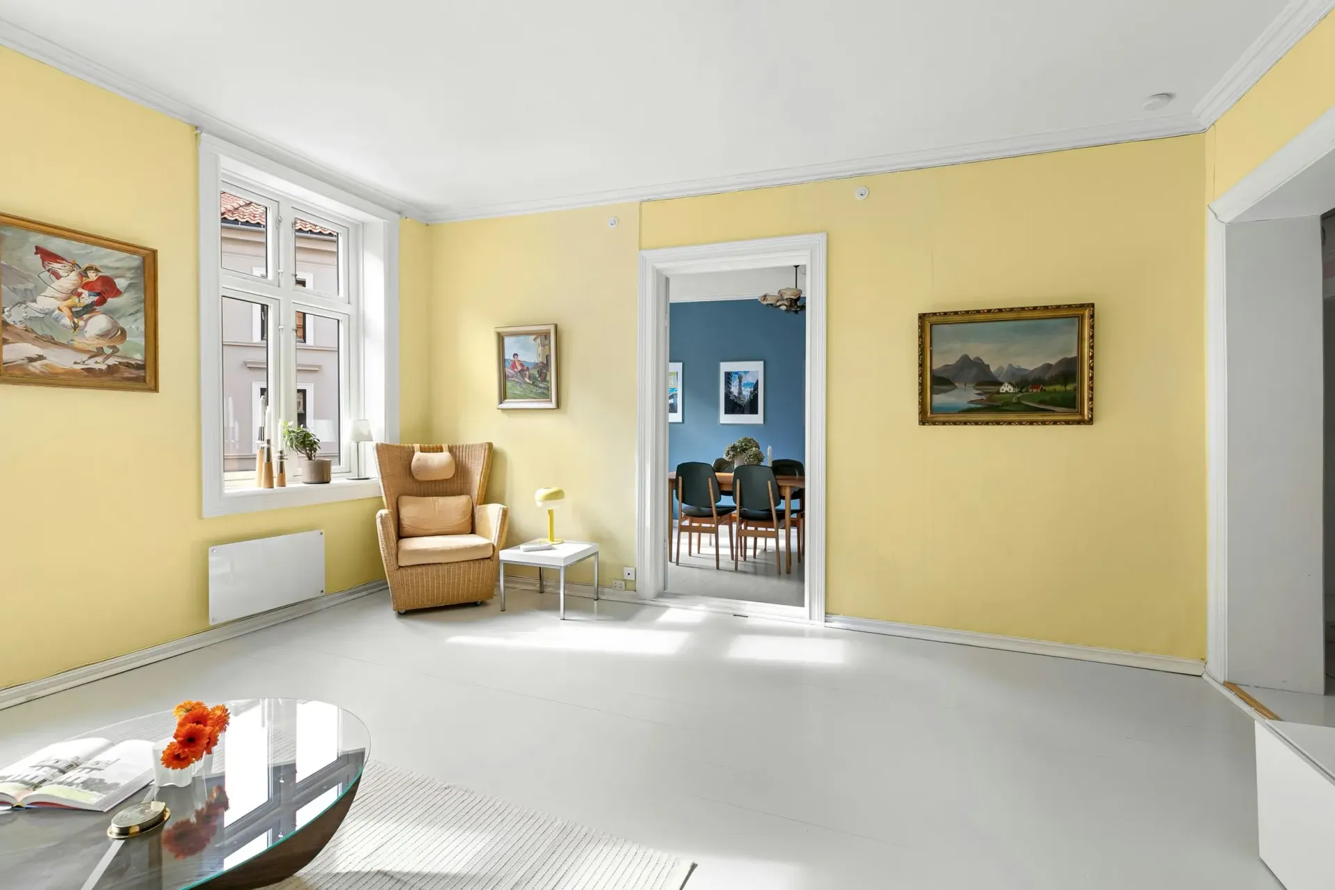 Bright living room with yellow walls, white floor, tan armchair, and framed paintings.