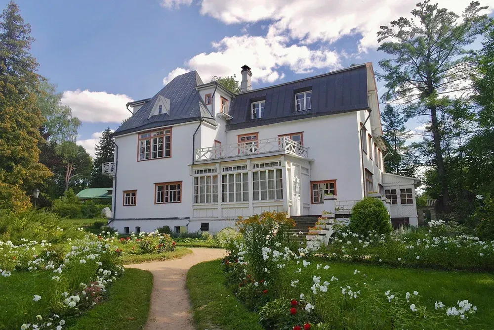 Large white country house with a glass sunroom, surrounded by a lush garden and trees under a blue sky.