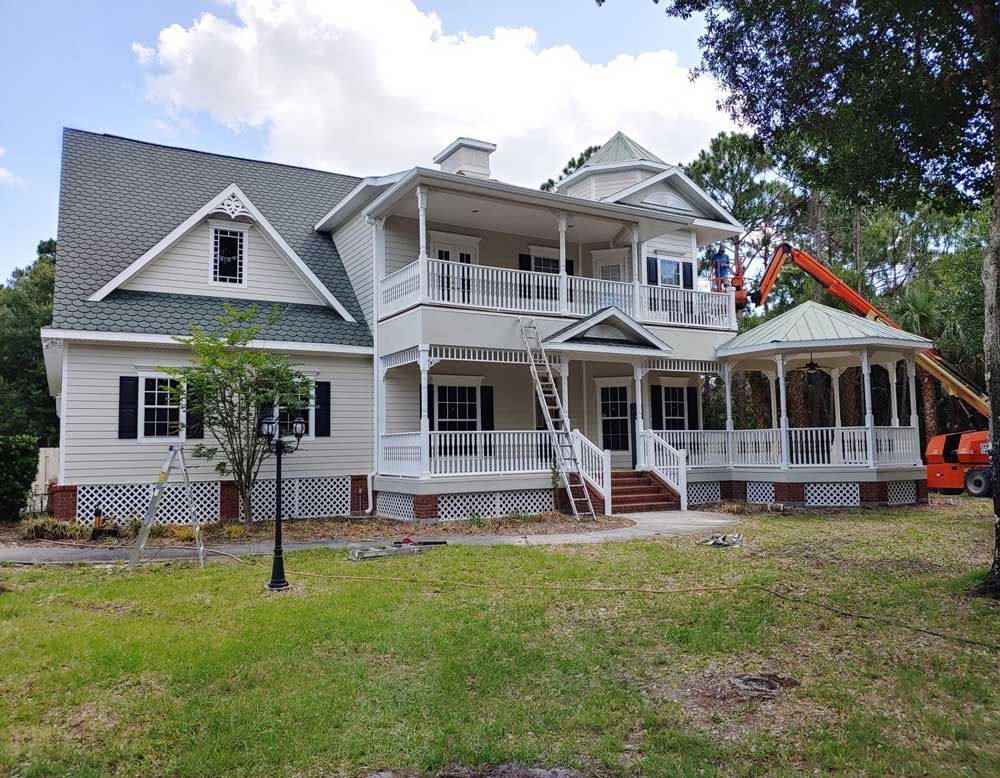 Large gray Victorian-style house with white trim, wraparound porch, and a small excavator on the right.