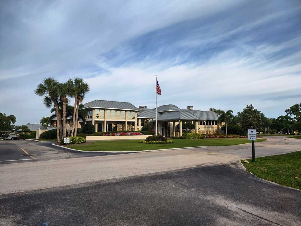 Large building with a flagpole, palm trees, and a parking lot under a cloudy sky.