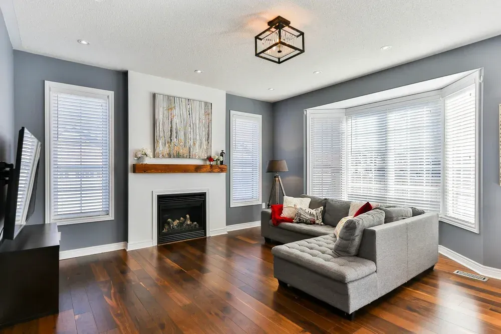 Bright modern living room with gray sectional, fireplace, hardwood floors, and large windows with white blinds