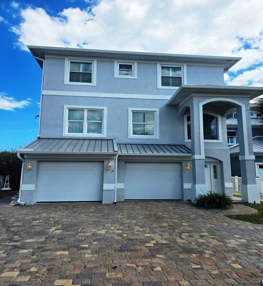 Three-story gray house with white trim, two garage doors, and a brick driveway under a blue sky
