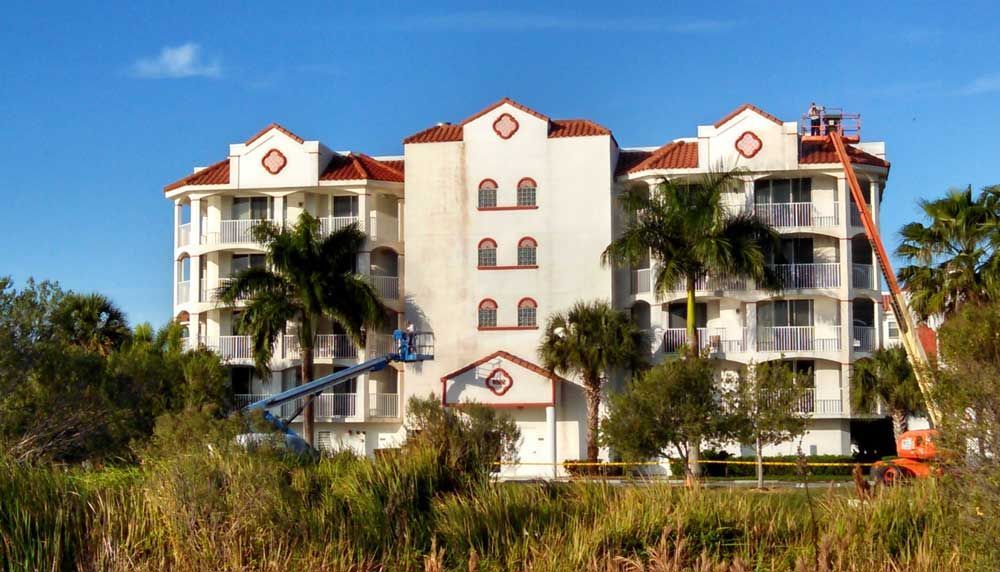 White beachfront condo with red roofs and balconies, partly obscured by palm trees under a clear blue sky