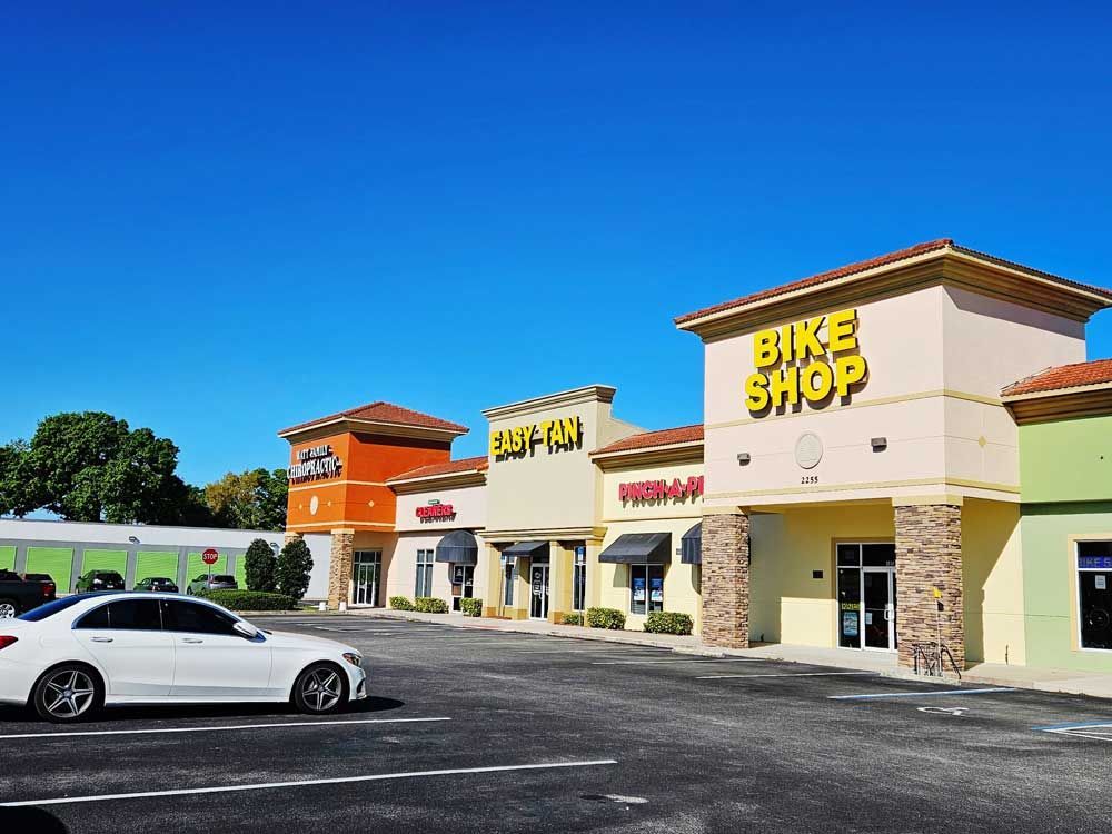 Shopping plaza with a bike shop, restaurant, and parked white car on a sunny day