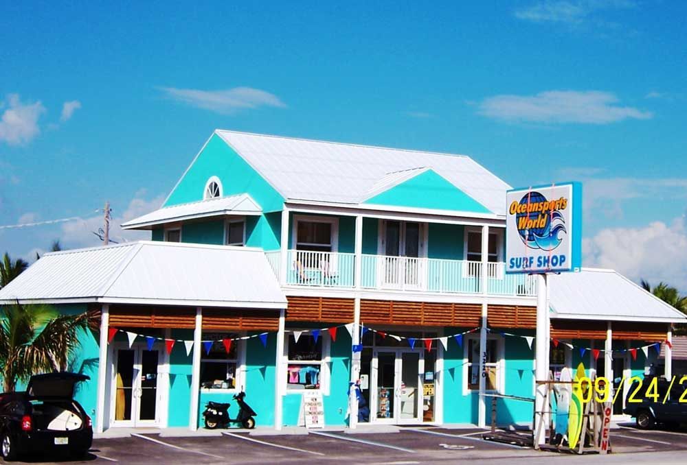 Turquoise beachside shop with white balconies, red awnings, and a parking lot under a clear blue sky