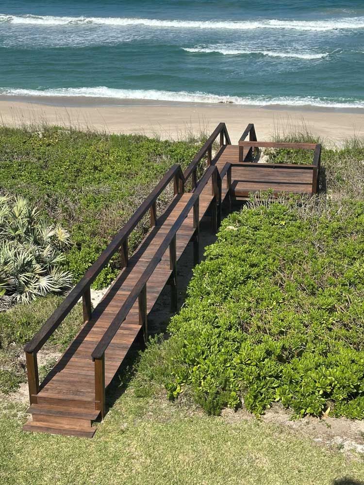 Wooden boardwalk with railings leading through green dunes to a sandy beach and ocean waves