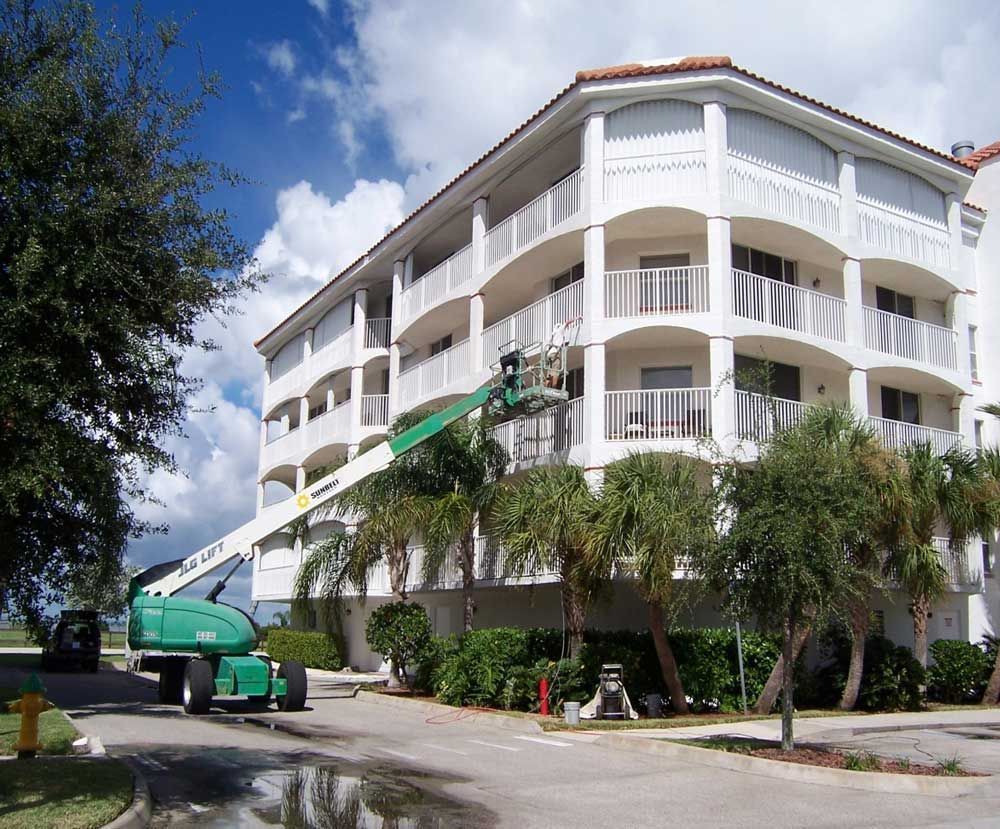 White multi-story building with balconies, a green cherry picker, and palm trees on a sunny street