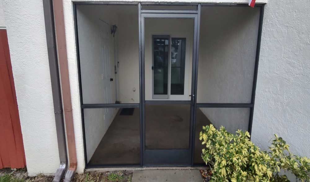 Entrance with screened double doors and a narrow walkway, framed by white walls and a small bush on the right.