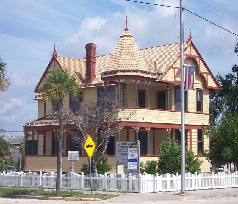 Victorian-style house with a wraparound porch and turret behind a white picket fence on a sunny street.