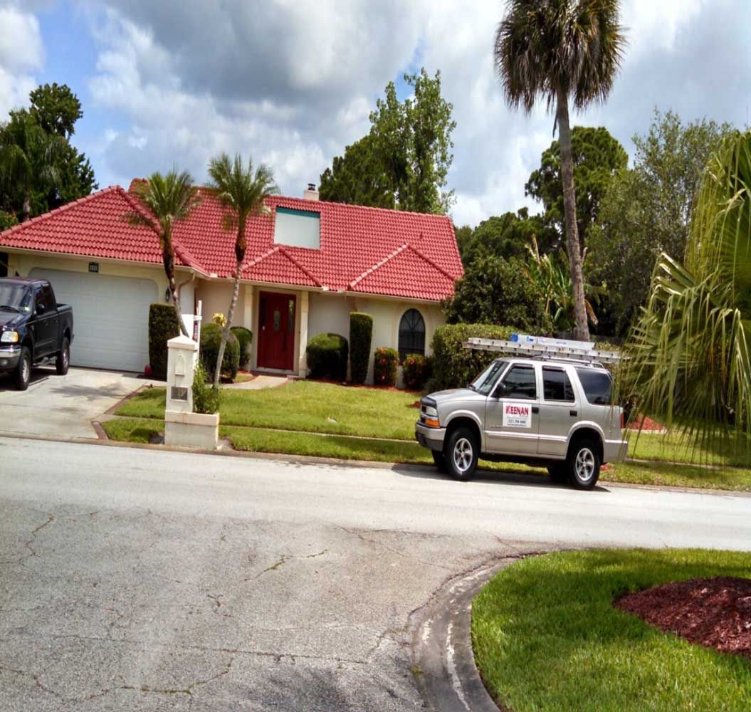 Suburban house with red roof, palm trees, and two parked vehicles on a driveway under a cloudy sky