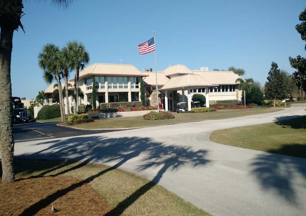 Large two-story suburban building with an American flag, palm trees, and a curved driveway under a clear blue sky
