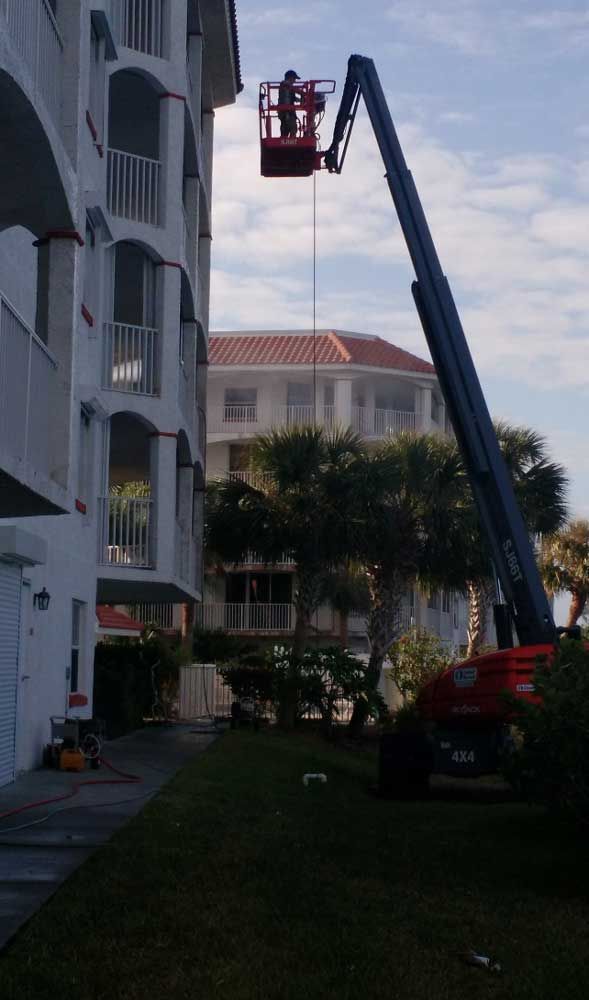 Worker in red lift basket inspecting a building exterior next to a palm tree at dusk