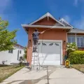 Person on a ladder painting the front of a tan house with a white garage door