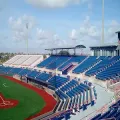 Empty sports stadium with blue and white seats beside a red running track under a partly cloudy sky