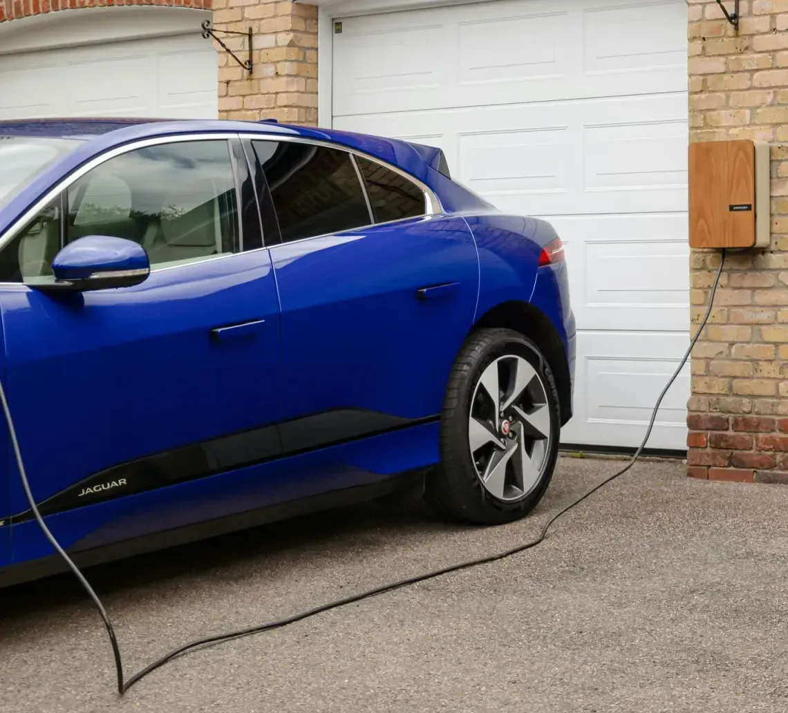 A blue electric car parked in a driveway, plugged into a wall-mounted charging station on a brick garage.