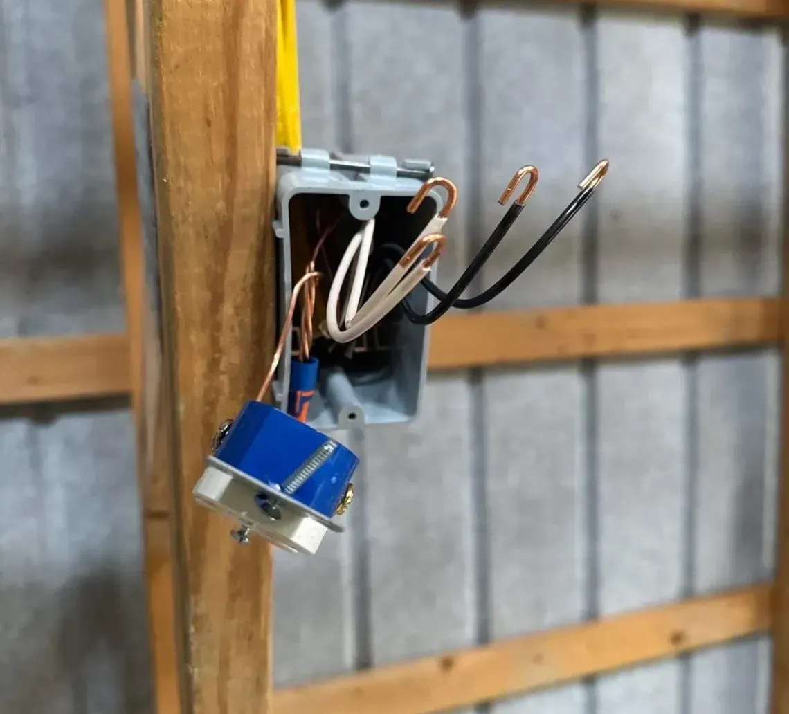 A gray electrical box attached to a wooden stud with exposed white, black, and copper wiring and a blue wall switch.