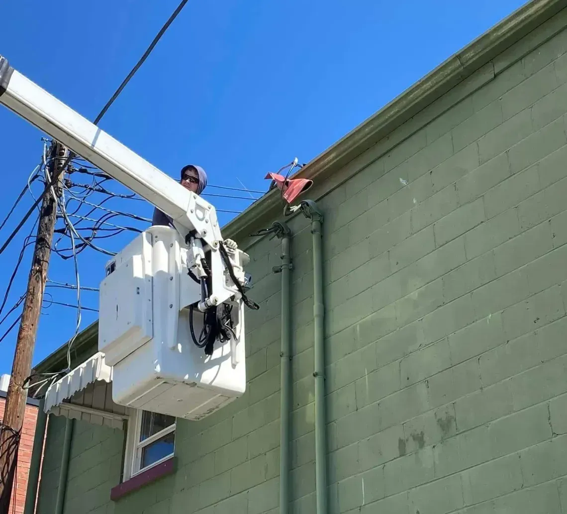 A worker in a white bucket truck boom reaches toward an electrical line attached to a light green brick building.