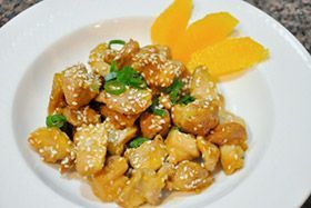 Glazed chicken pieces garnished with sesame seeds and green herbs on a white plate with mango slices