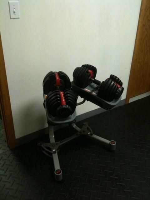 Adjustable dumbbell rack with black and red weights in an office corner.