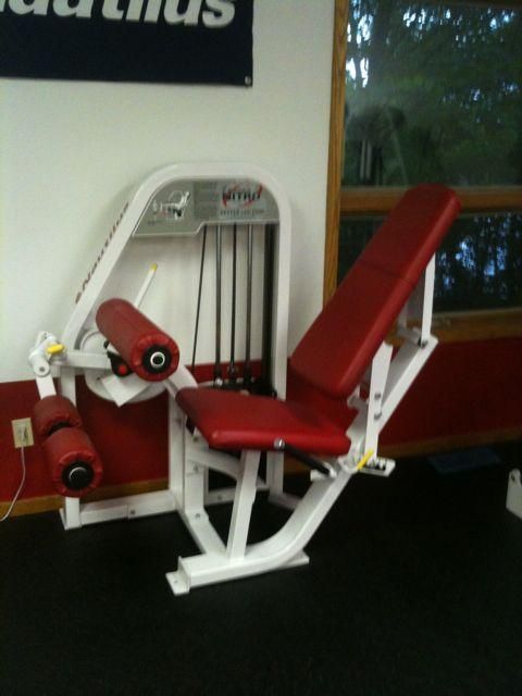 Red seated exercise machine with leg rollers in a gym room near a window.