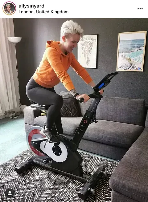 Person on a stationary bike in a living room, wearing an orange top and black leggings.