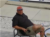 Man sitting in a black chair outdoors on a paved surface, wearing a cap and striped shirt, smiling.