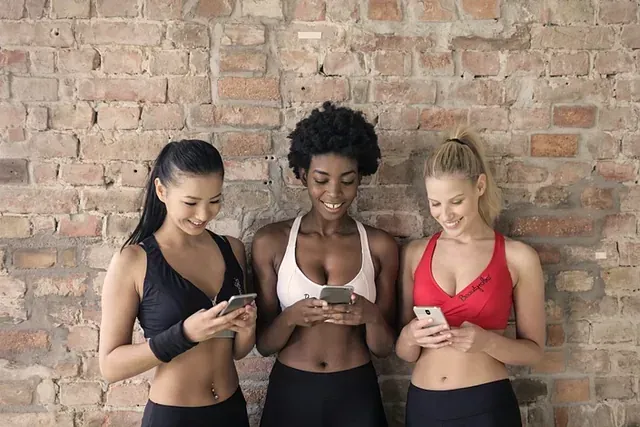 Three people in workout clothes checking phones in front of a brick wall