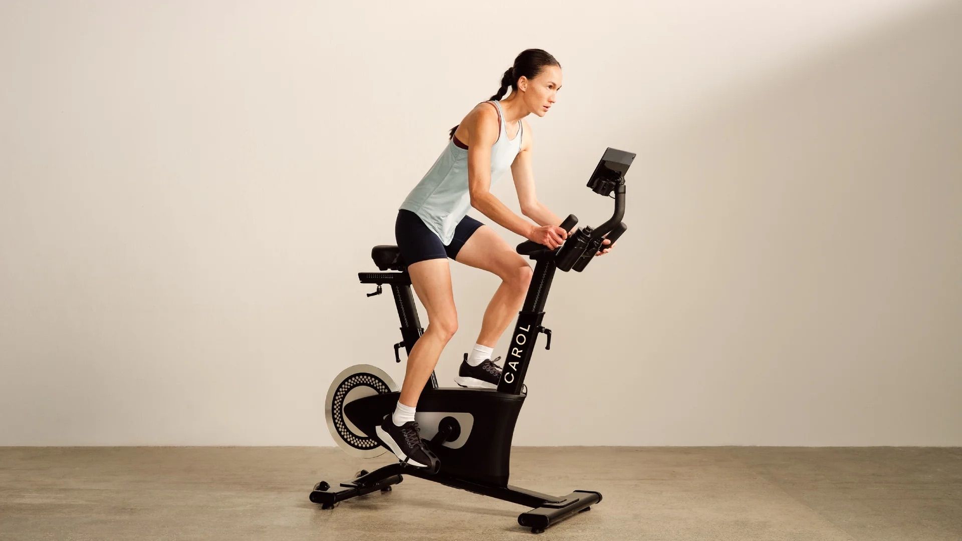 Woman exercising on a stationary bike in a minimalist studio, wearing a white top and blue shorts.