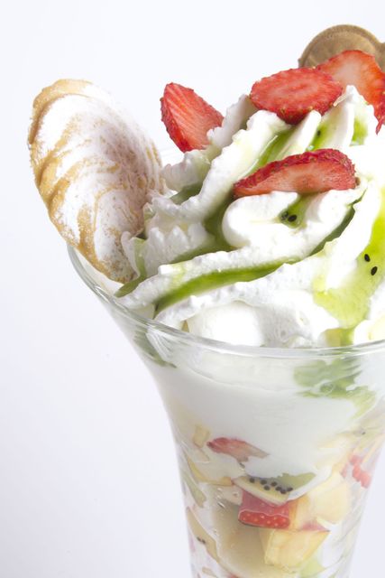 Una coppa di frutta in un bicchiere alto, guarnita con panna montata, fette di fragole, salsa al kiwi e un biscotto di meringa.