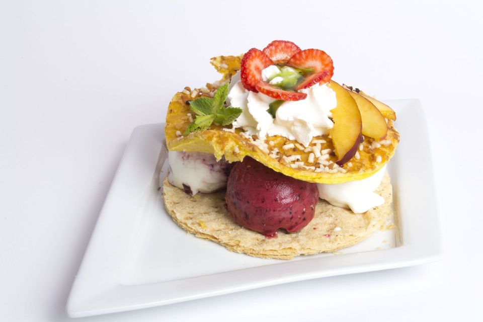 Un dessert elaborato con gelato, frutta, panna montata e sottili strati di pasta sfoglia su un piatto quadrato bianco.
