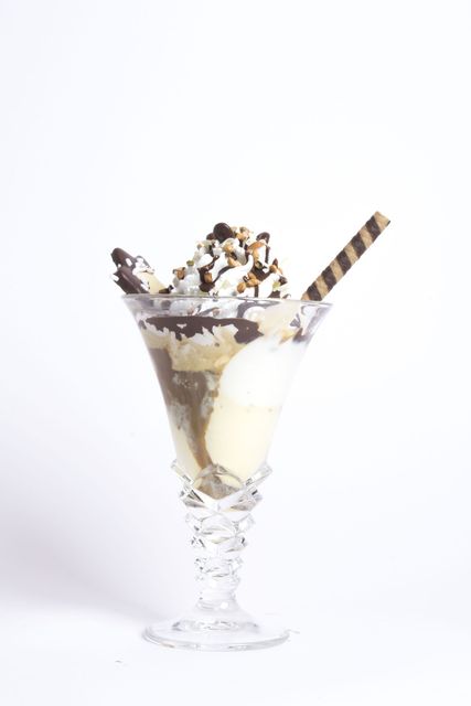 Una coppa di gelato in un bicchiere di cristallo, guarnita con panna montata, salsa al cioccolato, granella colorata e una cialda a righe, su uno sfondo bianco.