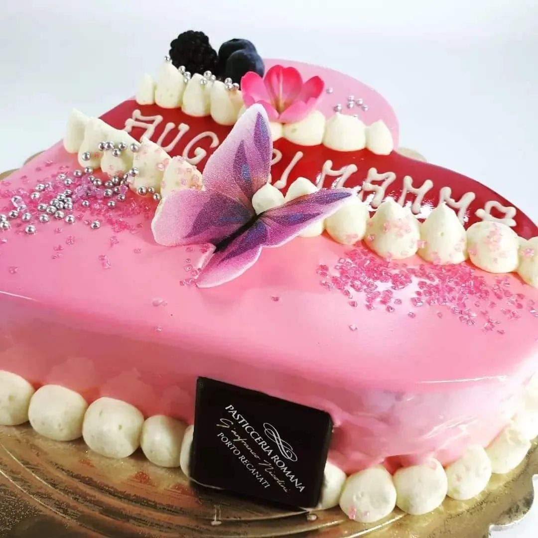 Una torta a forma di cuore con glassa rosa, una farfalla viola in cima, frutti di bosco e puntini di glassa con la scritta