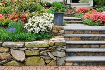 A stone wall with stairs leading up to a garden with flowers.