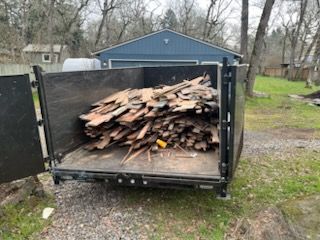 Open trailer loaded with stacked firewood in a gravel driveway beside a blue garage.