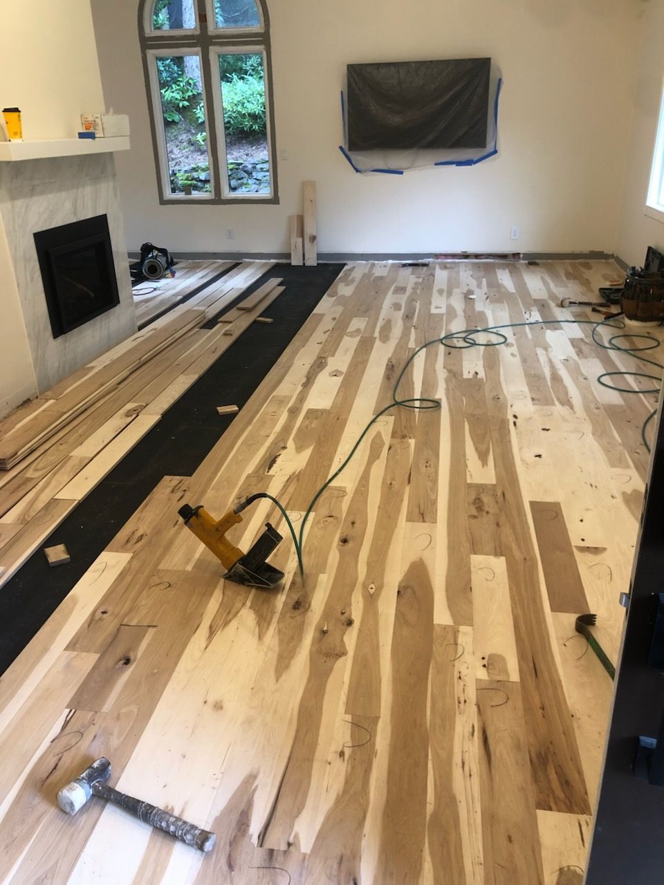 Partially refinished living room floor with tools, black inlay strips, fireplace, window, and wall-mounted TV.