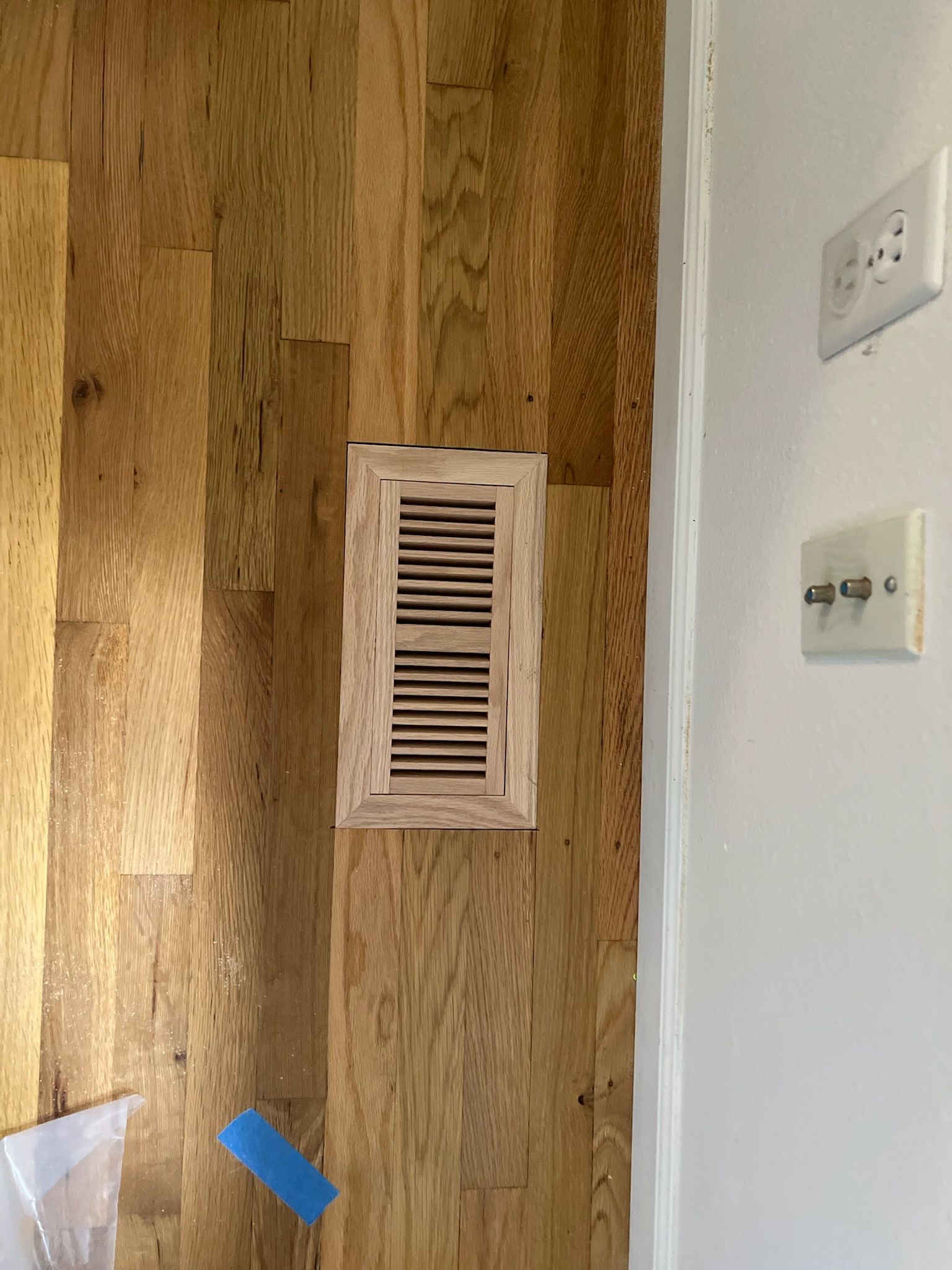Wood floor with a rectangular floor vent beside a white wall with electrical outlets