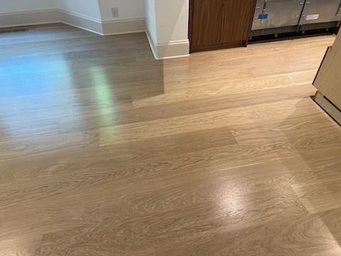 Light wood flooring in a modern room near a kitchen or hallway, with cabinets and sunlight reflections.
