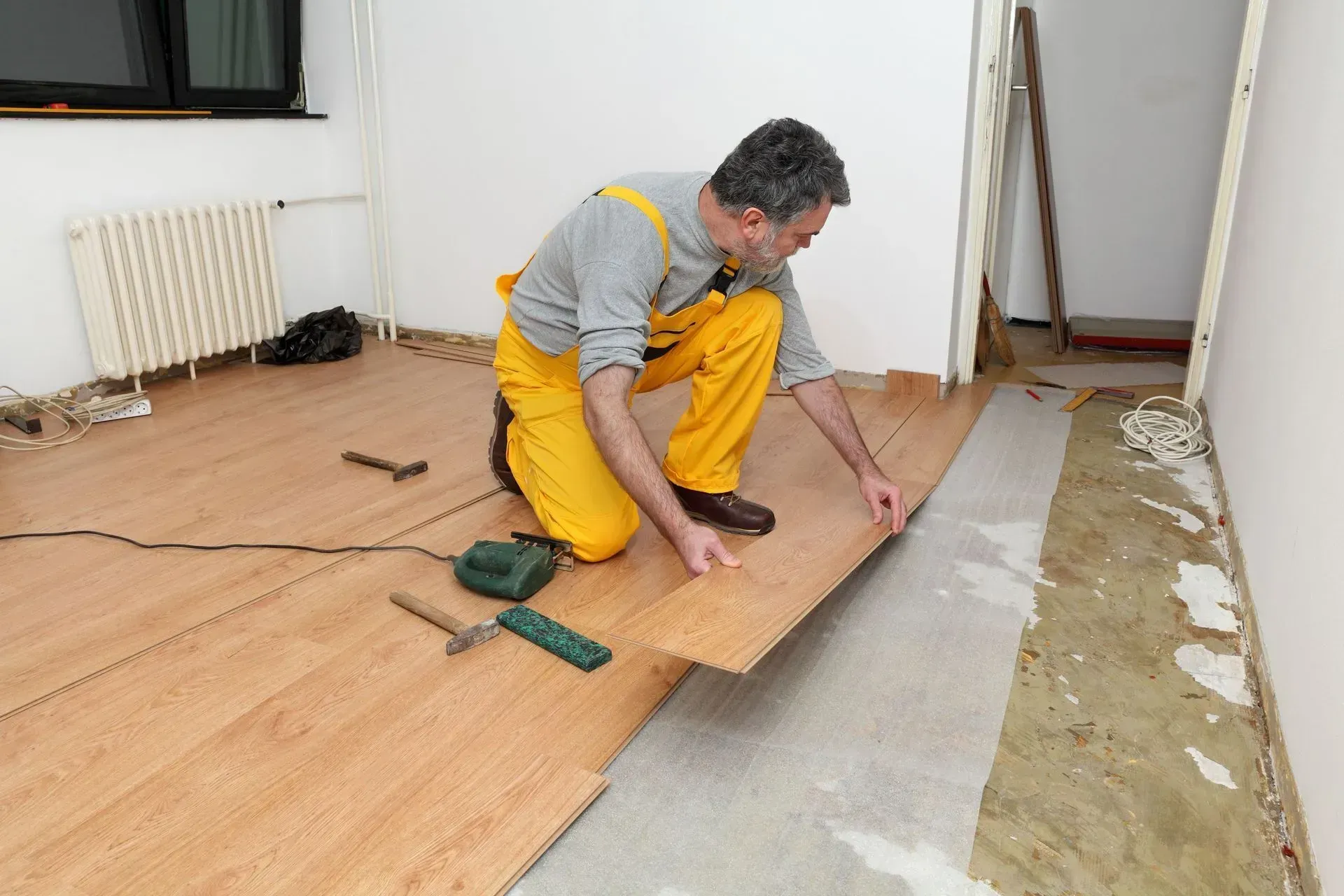 Man installing wooden flooring in a room, aligning a plank beside a radiator and exposed subfloor.