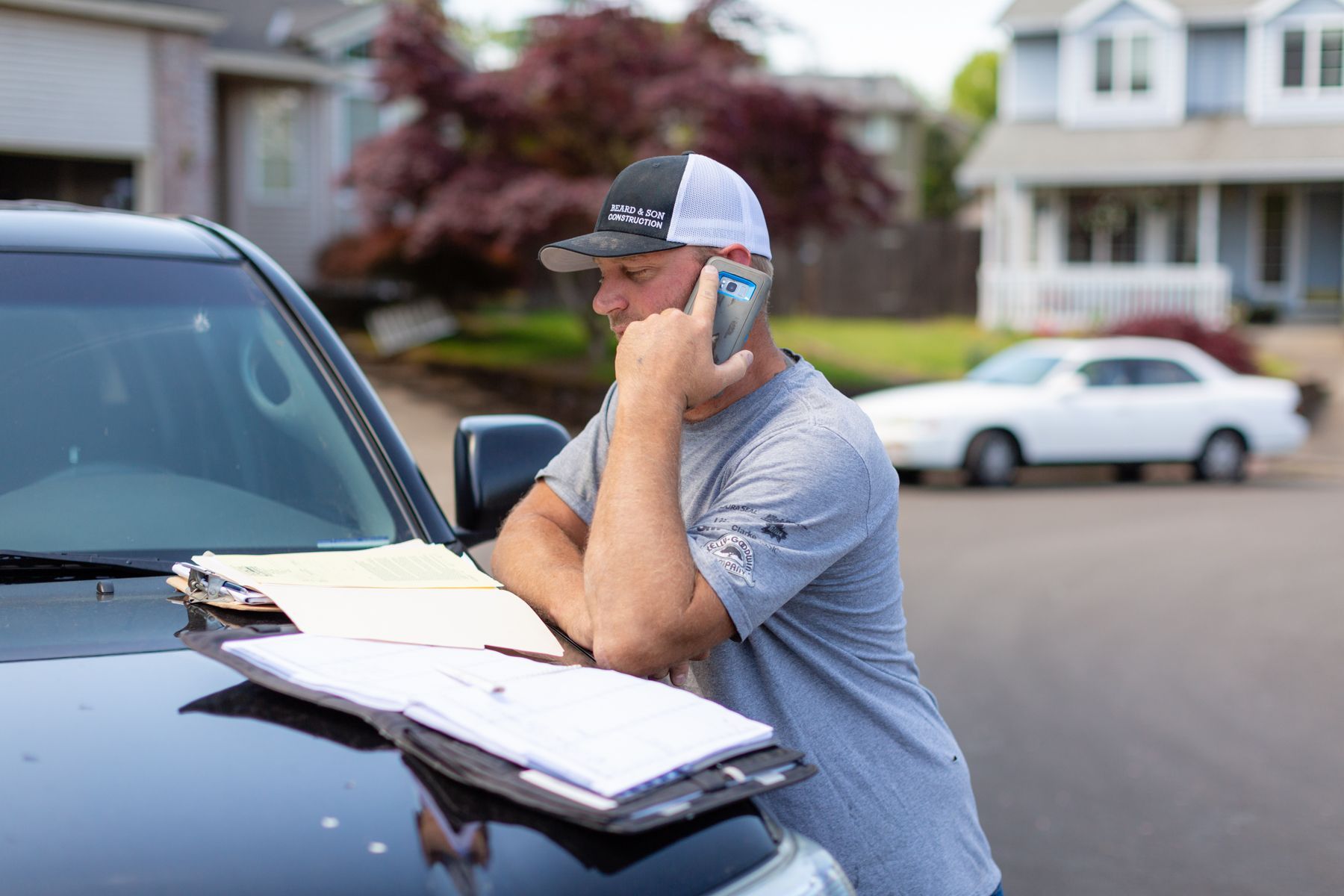 Man leaning on a car hood while talking on a phone, with papers spread on the hood, in a suburban street.